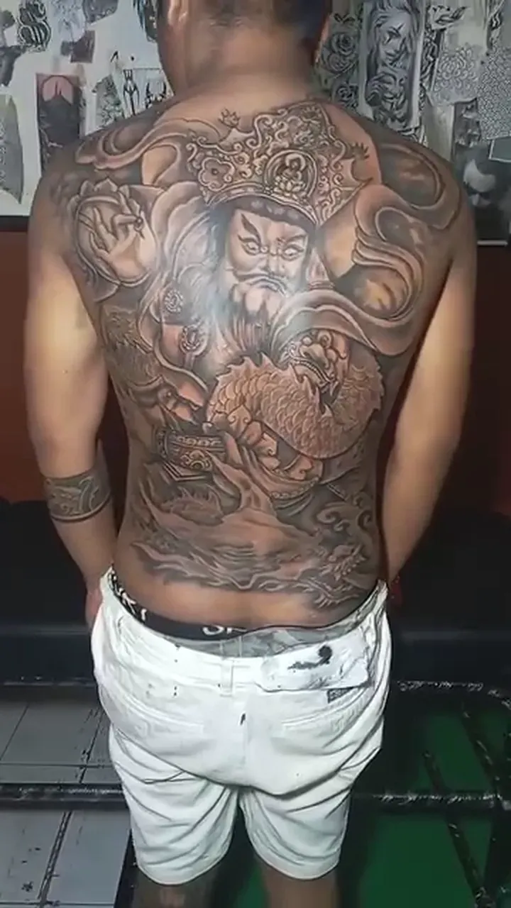 Large black and grey tattoo of a mythical figure with ornate headgear and clouds across the back
