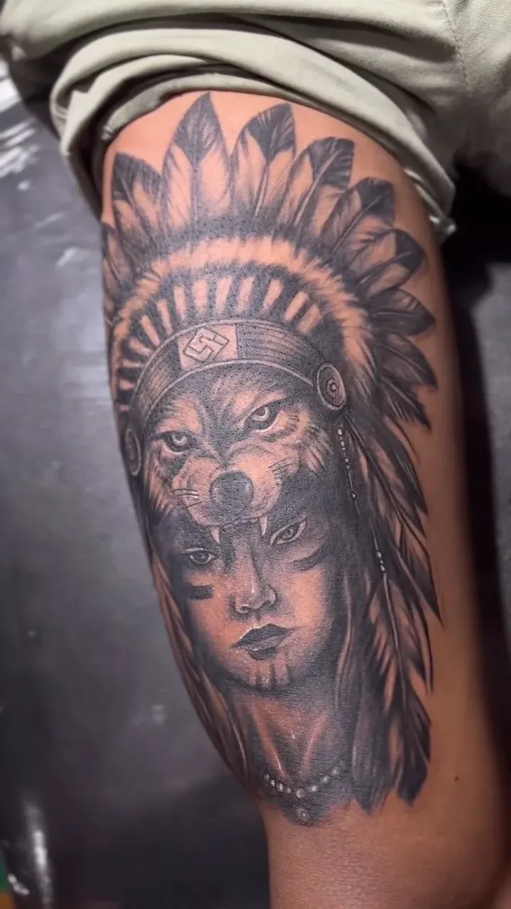 Black and grey realism fusion of a wolf and a warrior with a feather headdress, on a thigh